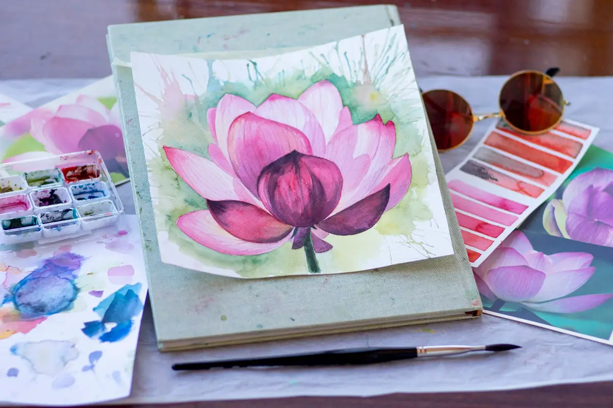 Aguarela de uma flor de lótus rosa sobre um caderno, junto a uma paleta e amostras de cor – Totenart